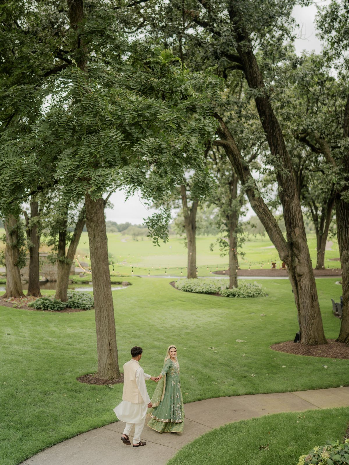Bride and groom in traditional Indian wedding attire walking on a park path.