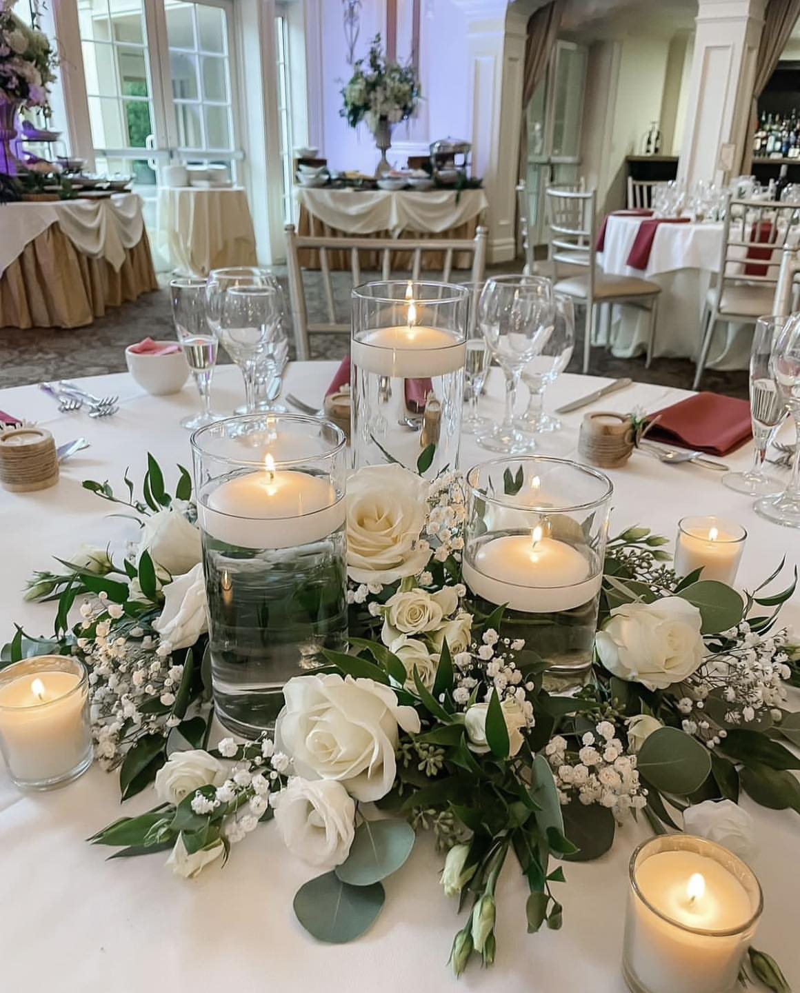 Floating white votive candles in glass cylinders surrounded by white roses and greenery on a table.