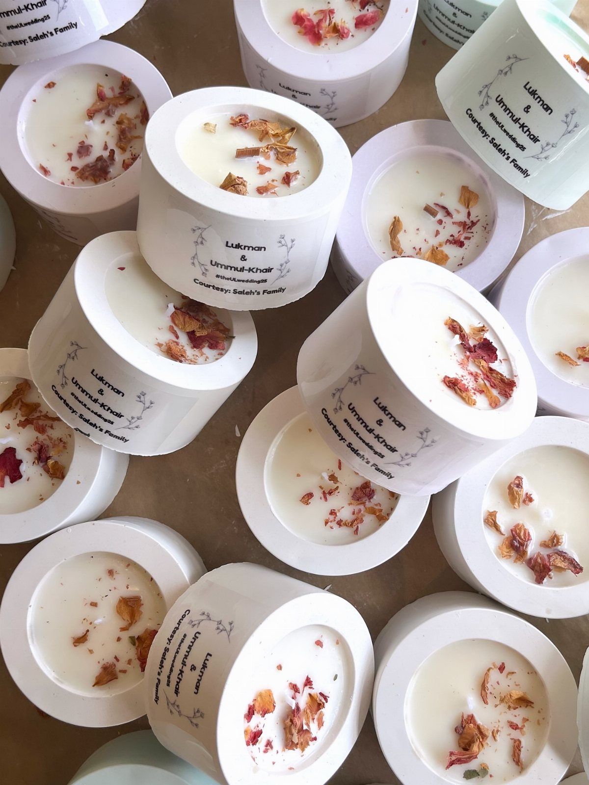 Numerous small white candles decorated with dried flower petals scattered on a light surface.