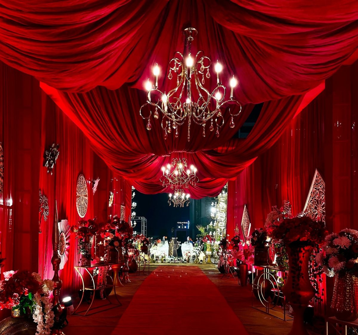 Red draped fabric ceiling with crystal chandeliers over a red carpet wedding aisle.