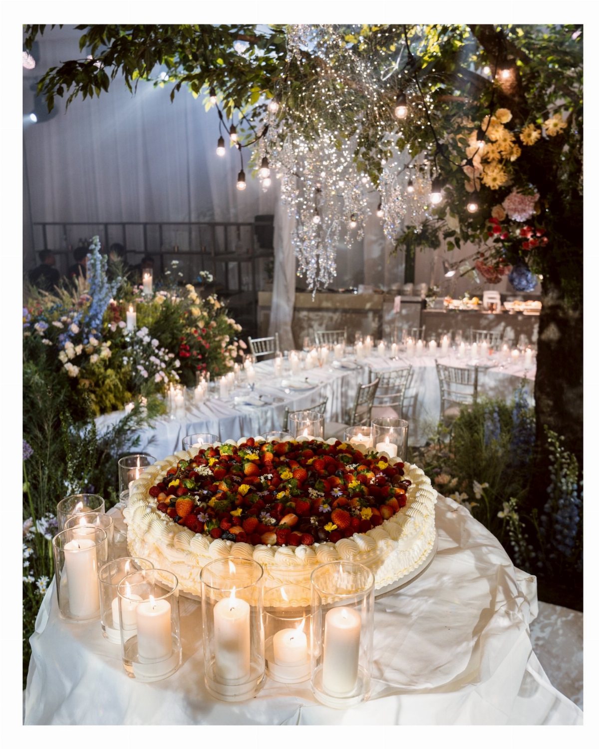 A berry-topped cake sits on a white table before a decorated wedding reception under fairy lights.