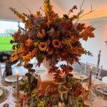 An autumnal floral centerpiece with orange roses and fall leaves on a decorated wedding reception table.