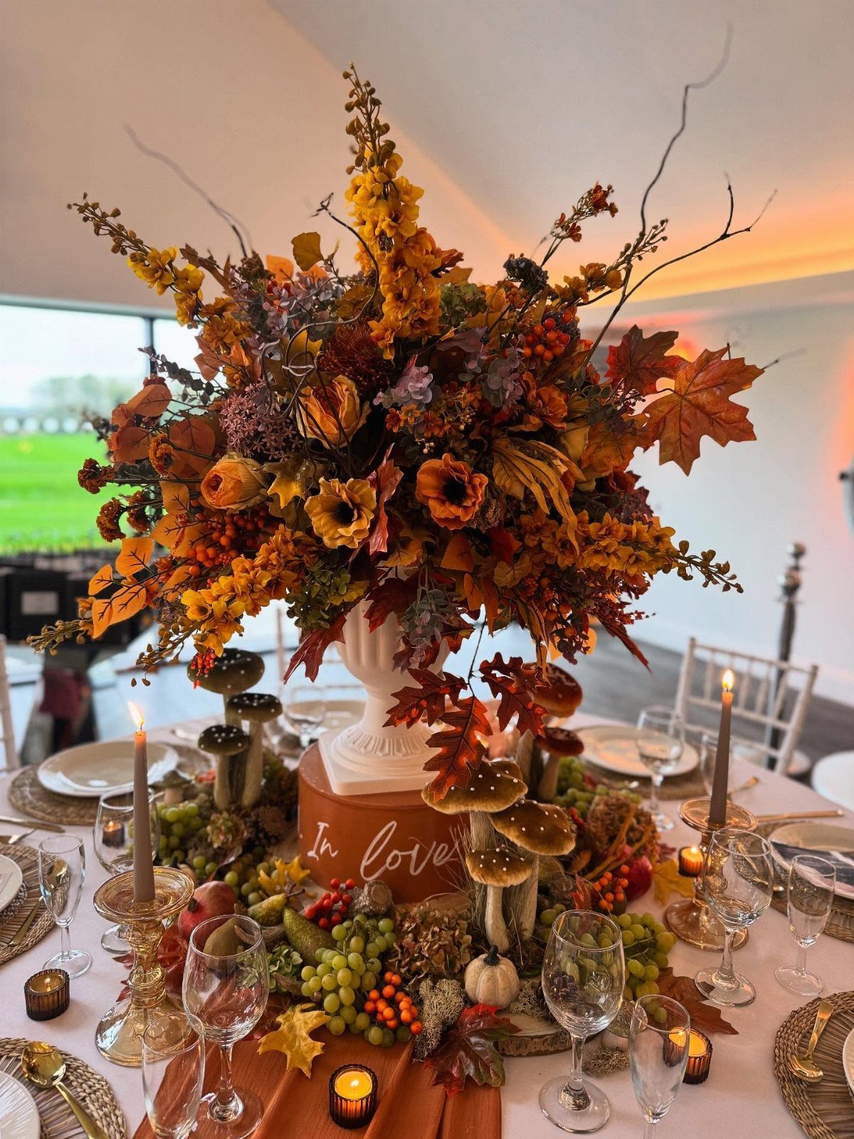 An autumnal floral centerpiece with orange roses and fall leaves on a decorated wedding reception table.