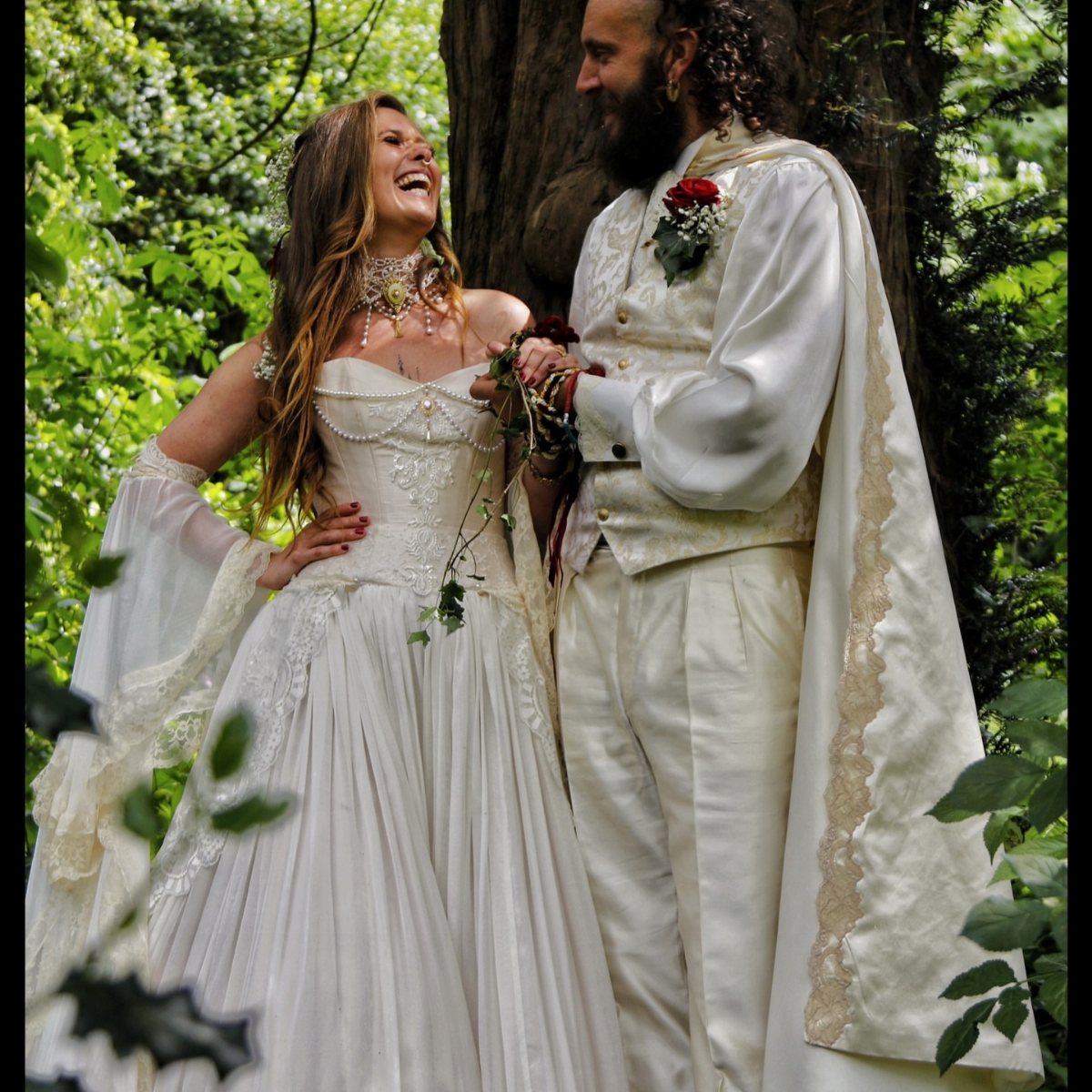 A couple in medieval inspired wedding attire smiling at each other in a lush green forest.