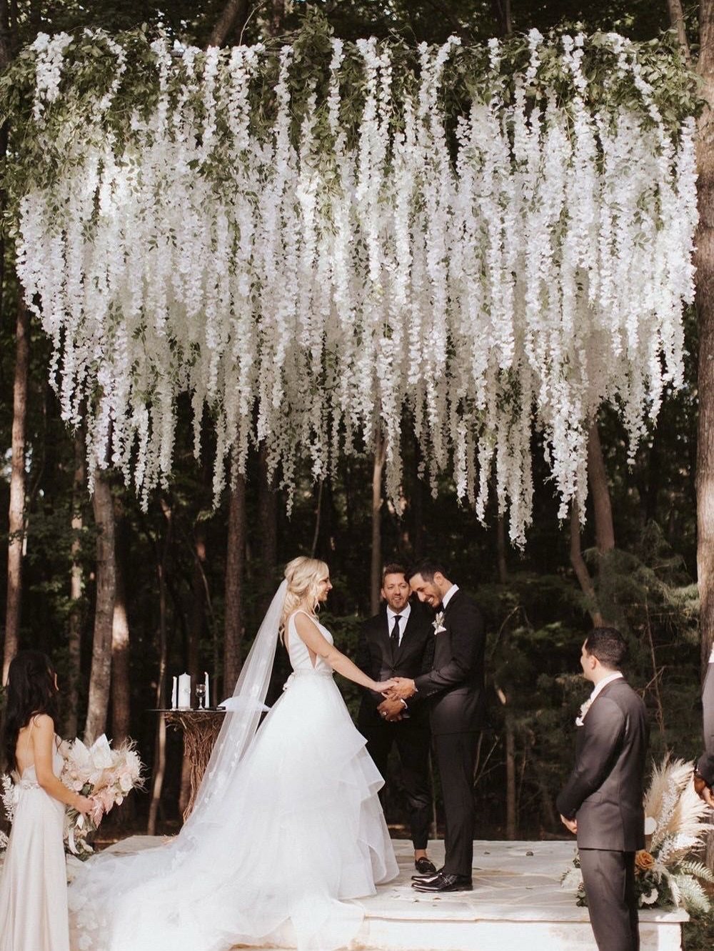 A bride and groom exchange vows under a large canopy of hanging white wisteria flowers in a forest.