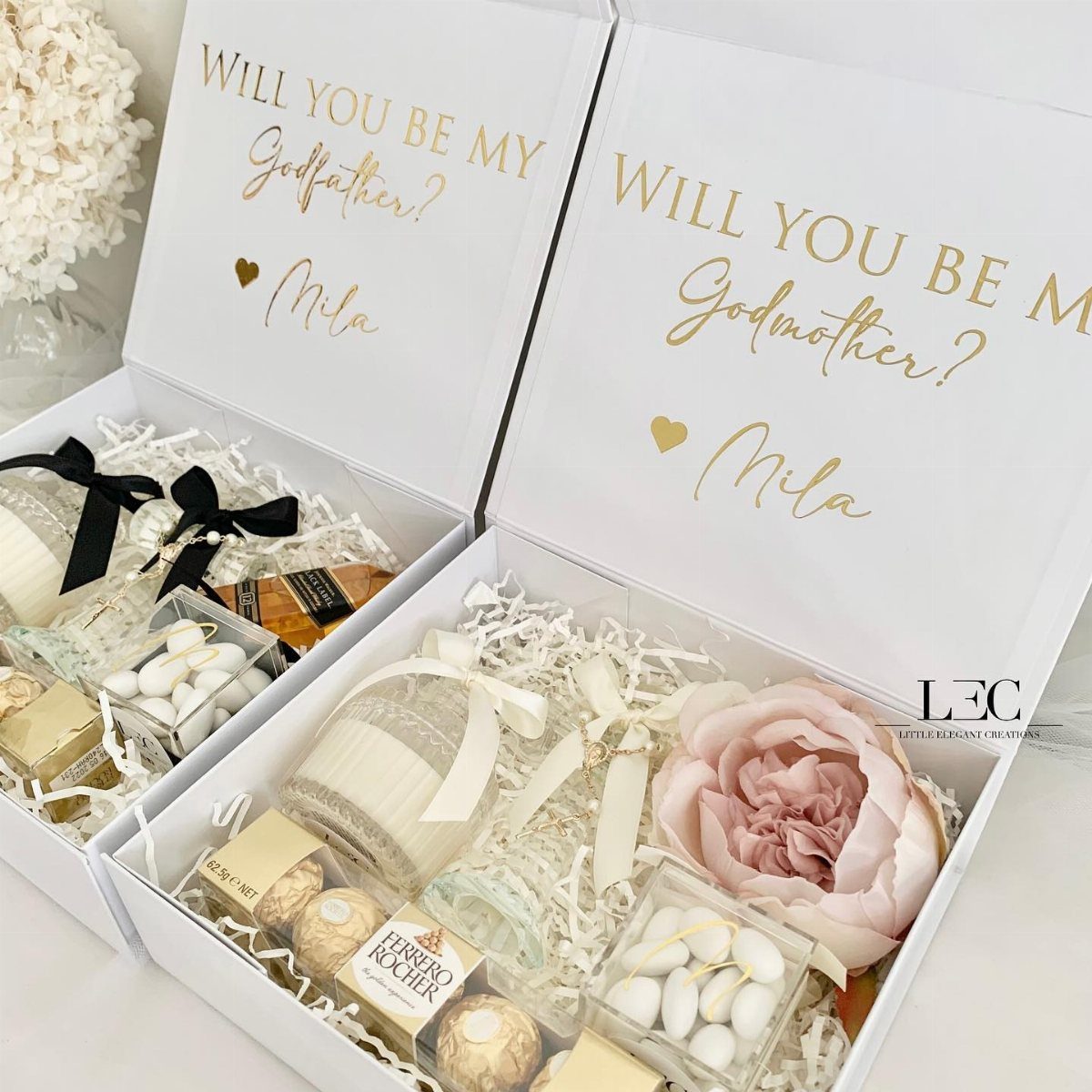 Two white godparent proposal boxes filled with chocolates, candles, and a pink flower.