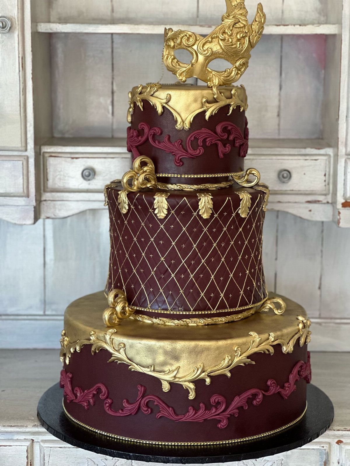 Three-tier burgundy and gold cake topped with a gold masquerade mask.
