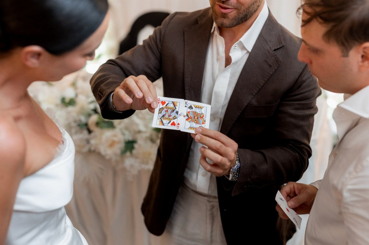 A man in a brown suit performs a card trick for a bride and a guest.