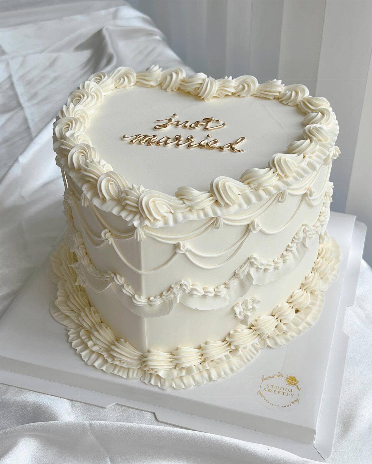 A heart-shaped white wedding cake with gold 'just married' lettering and cream piping.