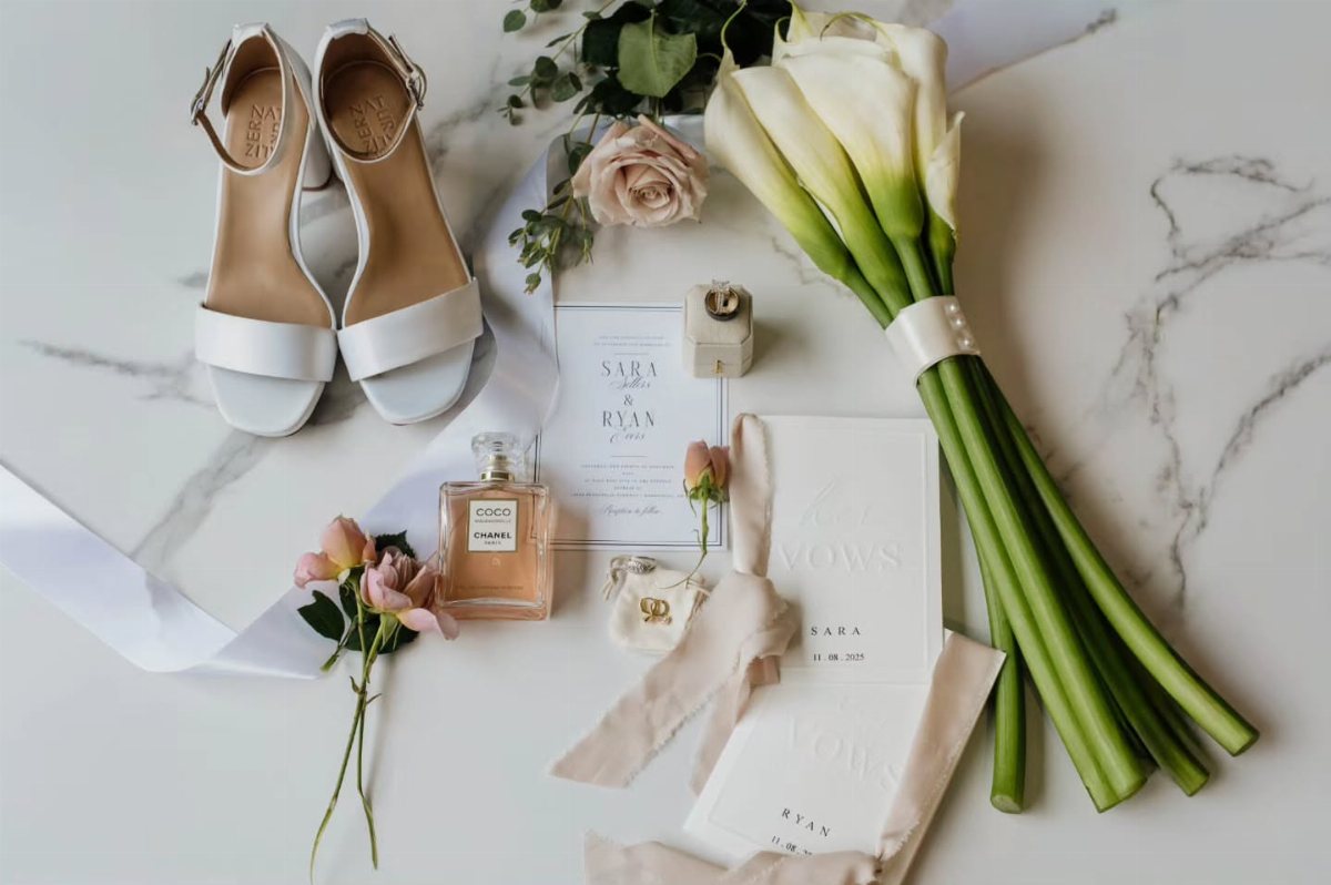 White wedding sandals, calla lilies, pink roses, perfume, and invitations arranged on a marble surface.