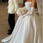 Bride in a shimmering lace and satin ballgown with detachable sleeves holding white flowers.