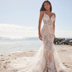 Woman in a tight white lace wedding dress posing on a sandy beach.
