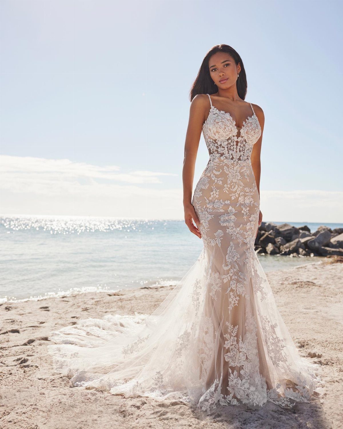 Woman in a tight white lace wedding dress posing on a sandy beach.