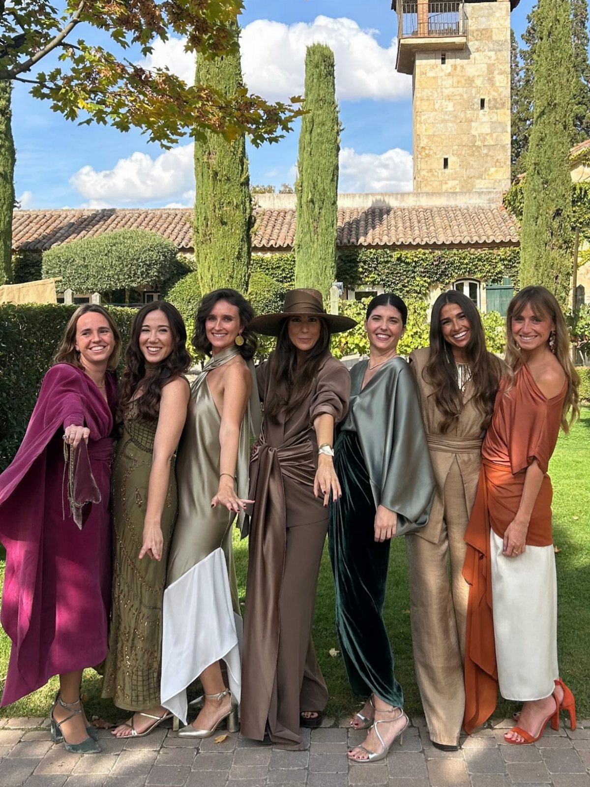Seven women in elegant formal attire posing together in a sunny outdoor garden setting.