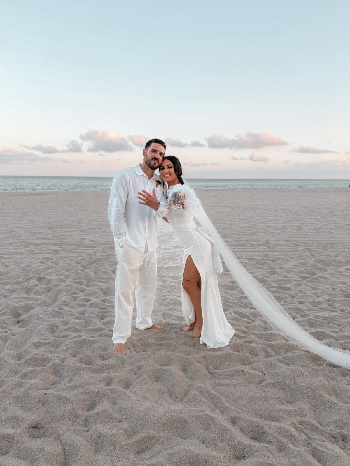 A bride and groom posing together on a sandy beach during sunset.