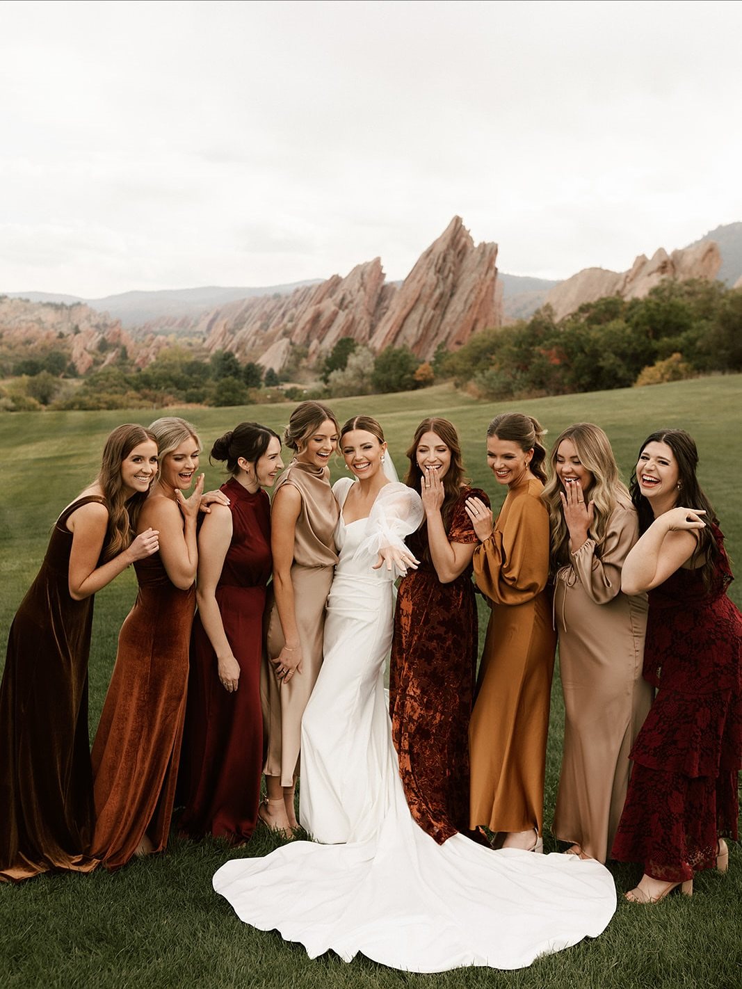 Bridesmaids in earthy-toned dresses pose with a bride in a grassy field near mountains.