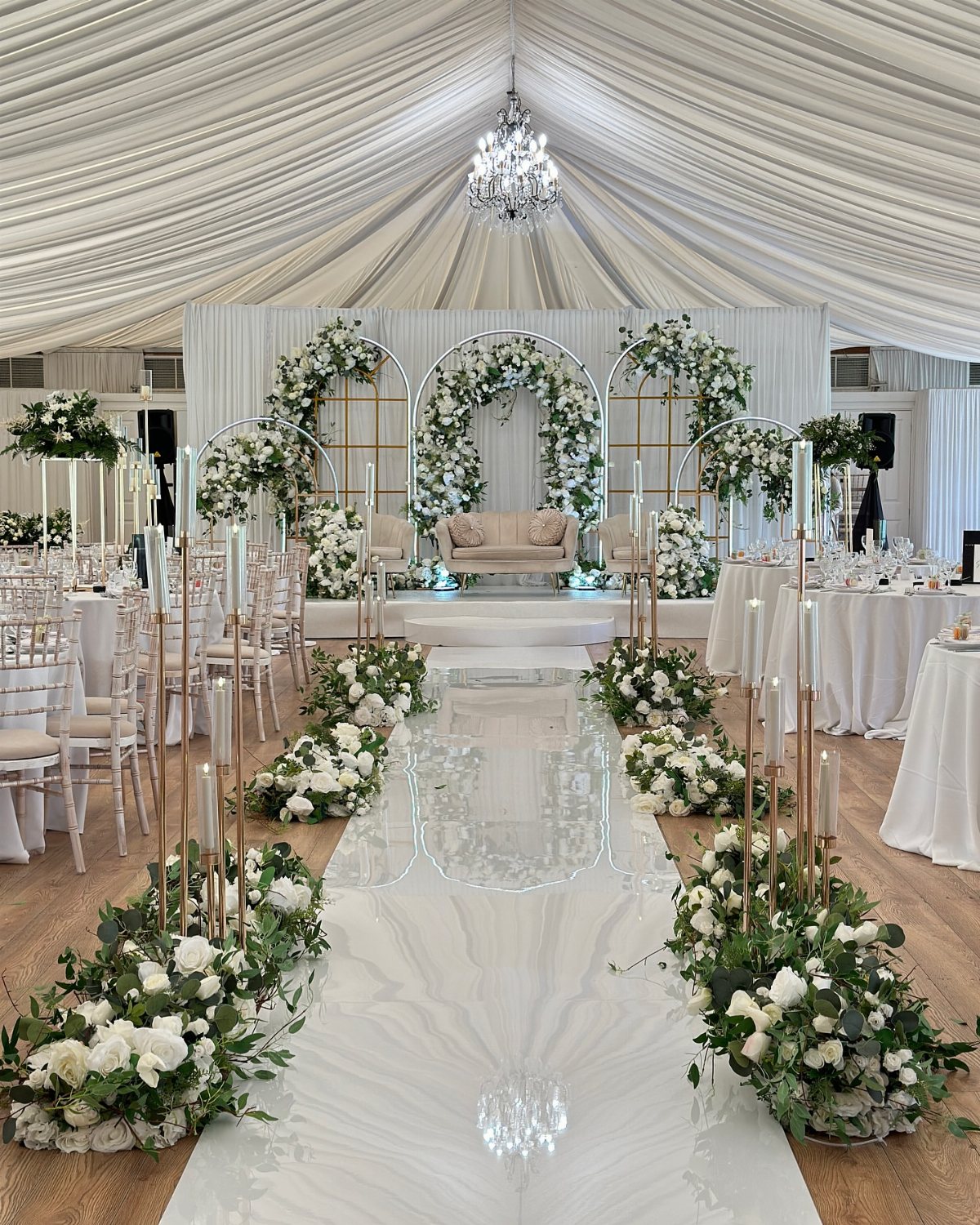 White wedding aisle lined with lush greenery and white floral arrangements under a draped tent.