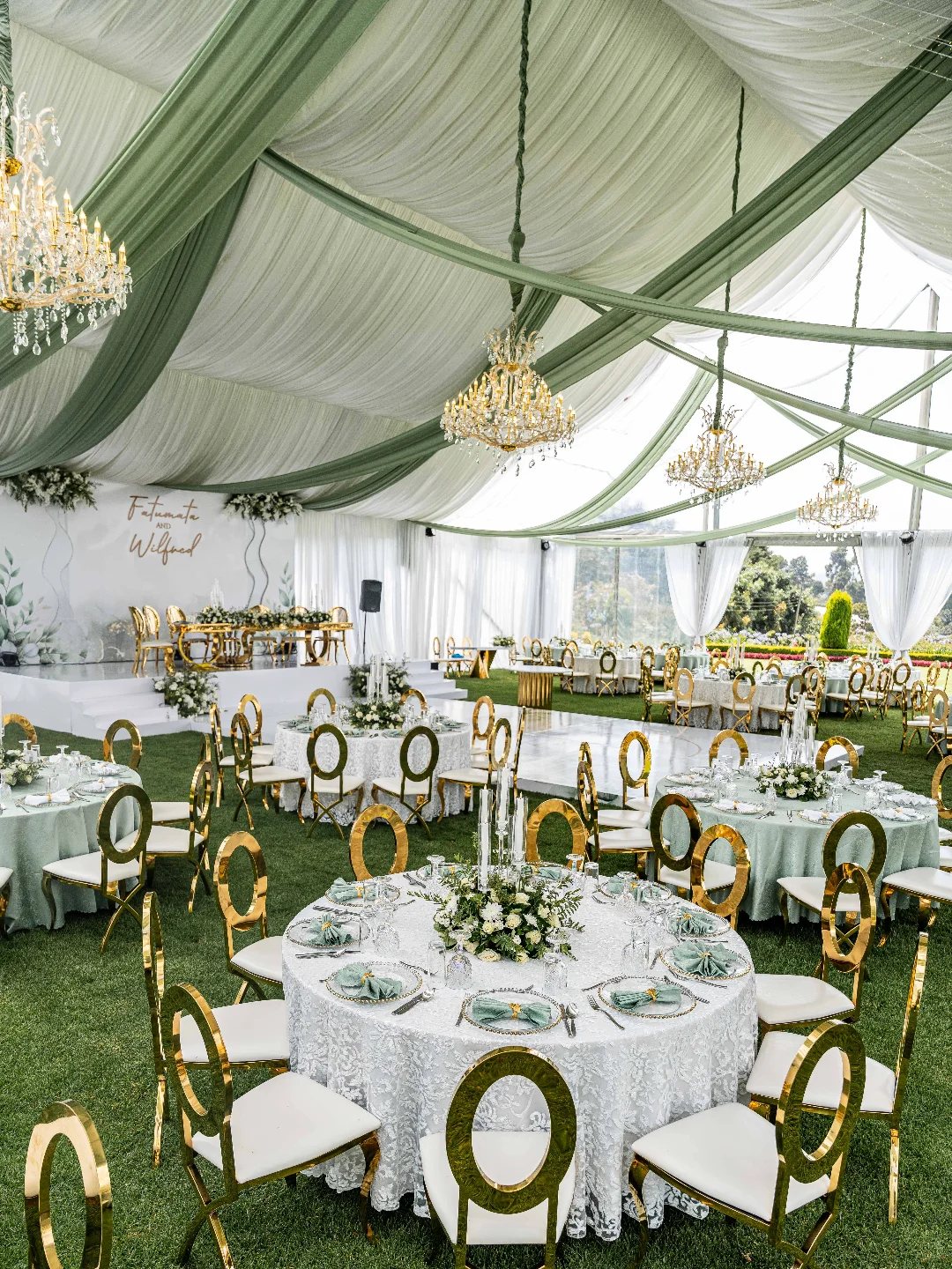 Wedding reception tables with white lace cloths and floral centerpieces set on grass under a draped tent.