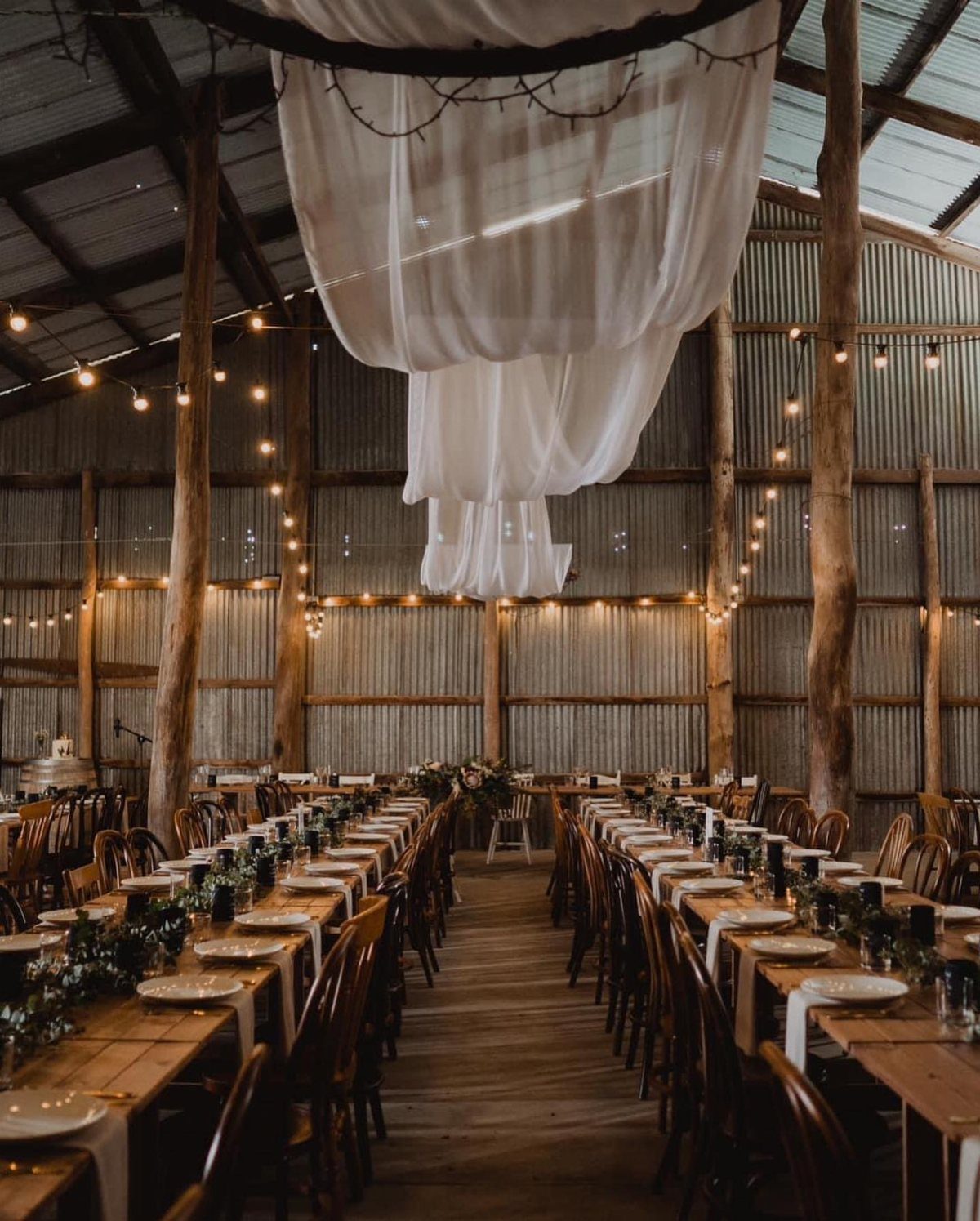 Rustic barn wedding venue with long wooden tables, white fabric draping, and string lights.