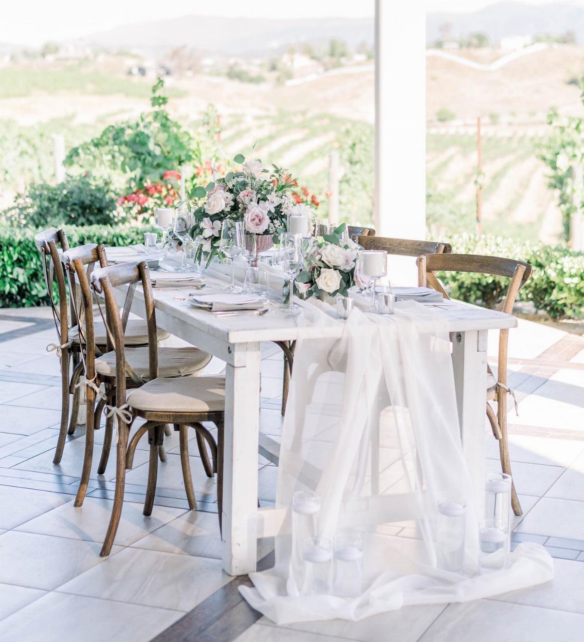 White wooden wedding table with pink floral centerpieces and wooden chairs outdoors.