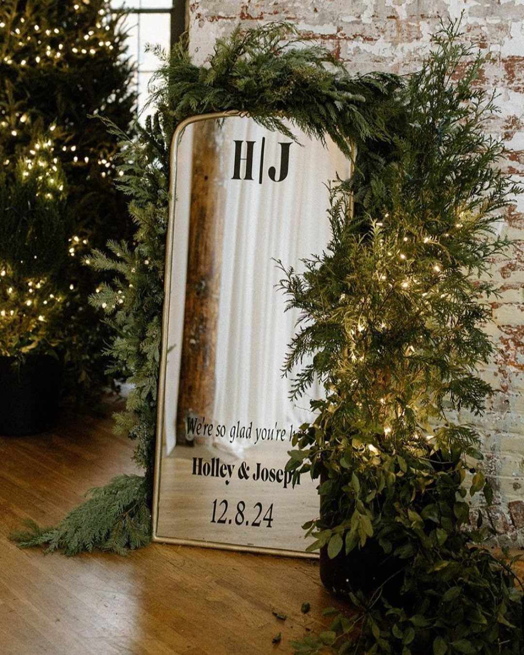 Arched wedding mirror framed with evergreen branches and fairy lights on wooden floor